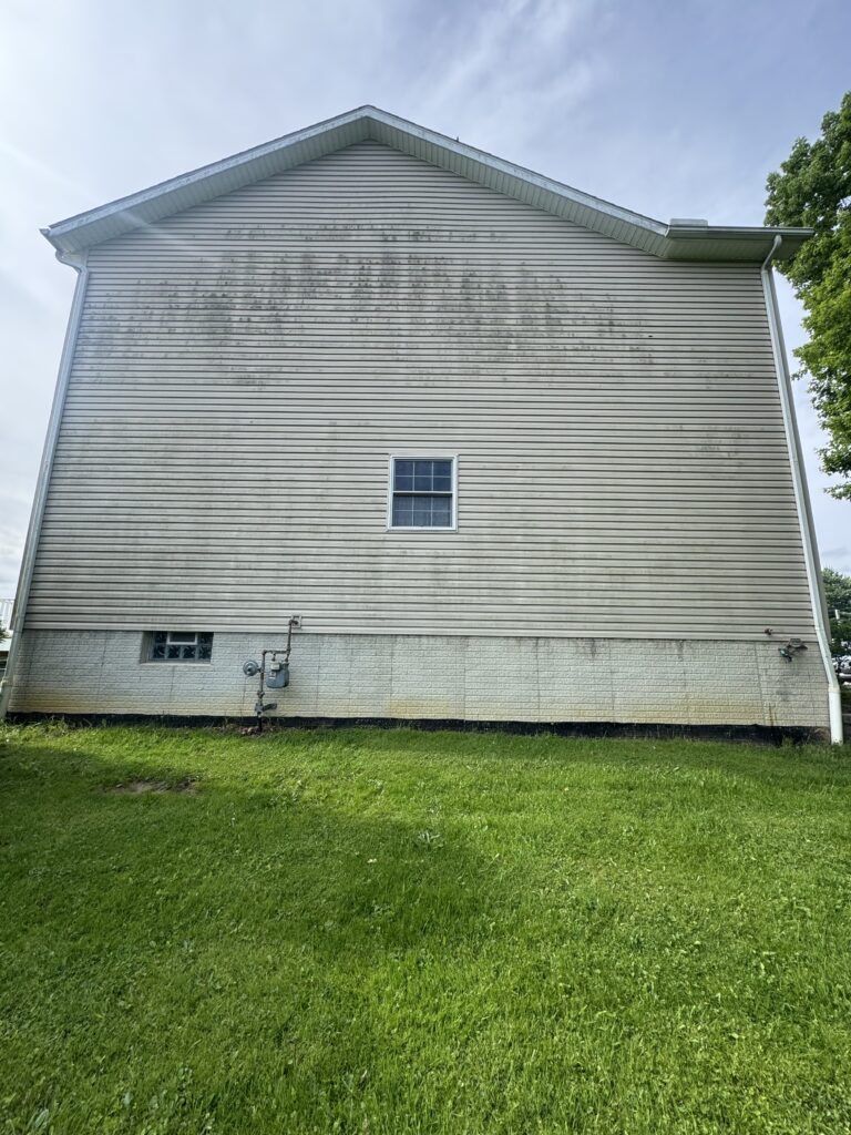 Side view of a beige vinyl-siding house wall with a small central window, utilities at the foundation, and a green lawn in the foreground.