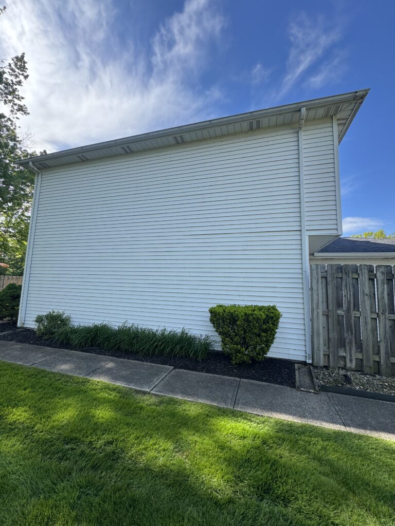 Side view of a white house with horizontal siding, gutter and downspout, small shrubs, mulch bed, sidewalk, and a wooden fence against a blue sky.