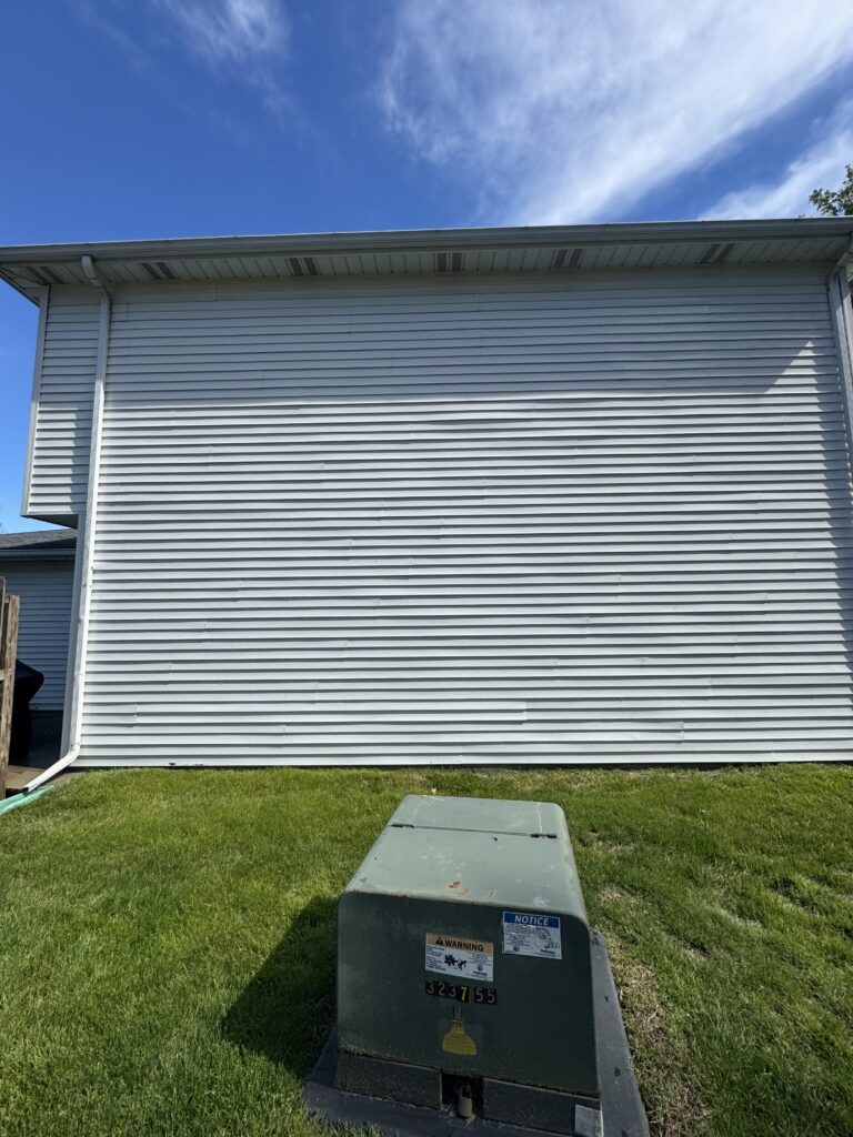 Backyard view of a white vinyl-sided house wall with a gray utility box in the grass and blue sky above.