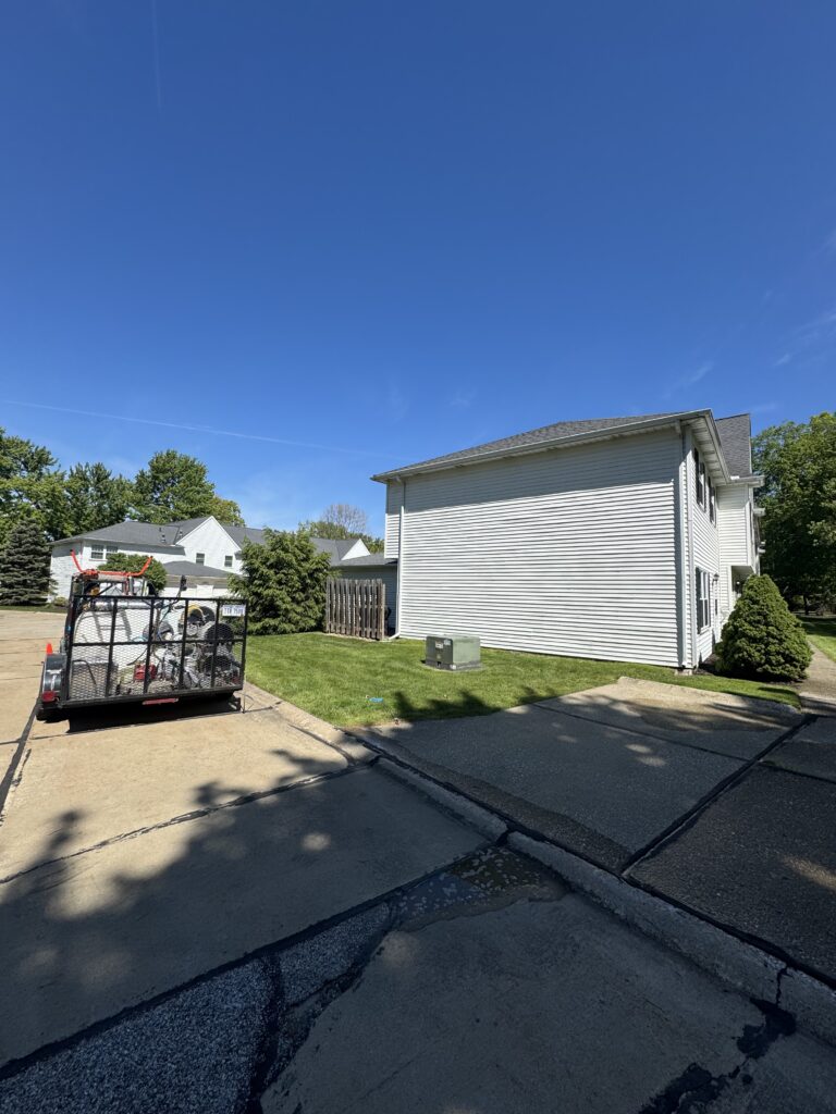 Suburban scene with a white two-story house, a trailer loaded with equipment in the driveway, and green lawns under a clear blue sky.