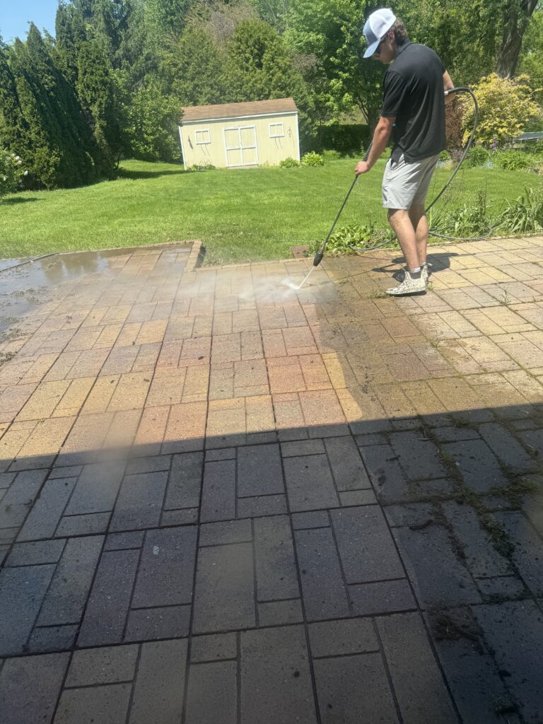 Man in a cap power-washing a brick patio with a high-pressure hose; spray plume visible, a grassy yard and a small shed in the background.