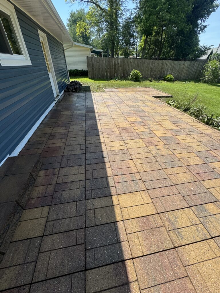 Backyard patio with brown and tan brick pavers beside a blue house, a wooden fence, and a grassy yard beyond.