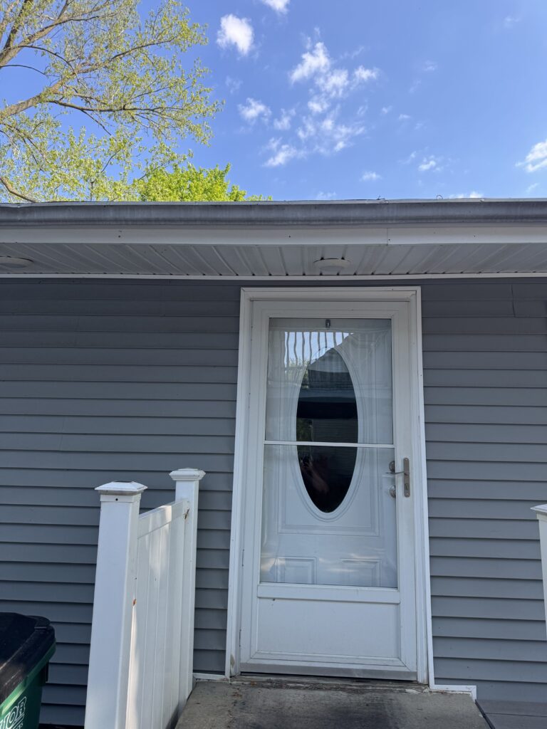 Front door with an oval glass panel and white trim on gray siding, a small porch, and a white railing in front of a blue sky.