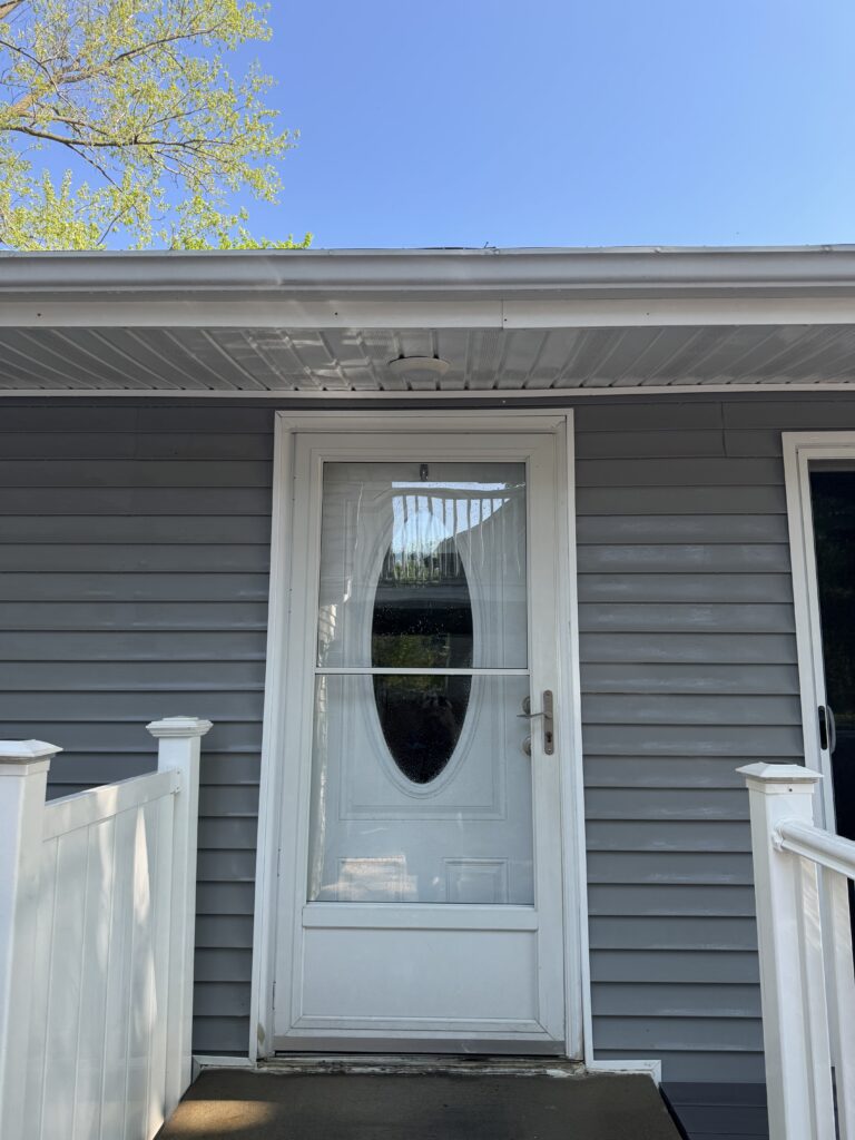 Front exterior view of a gray house with a white front door featuring an oval glass panel and white trim, flanked by white railing on a small porch.
