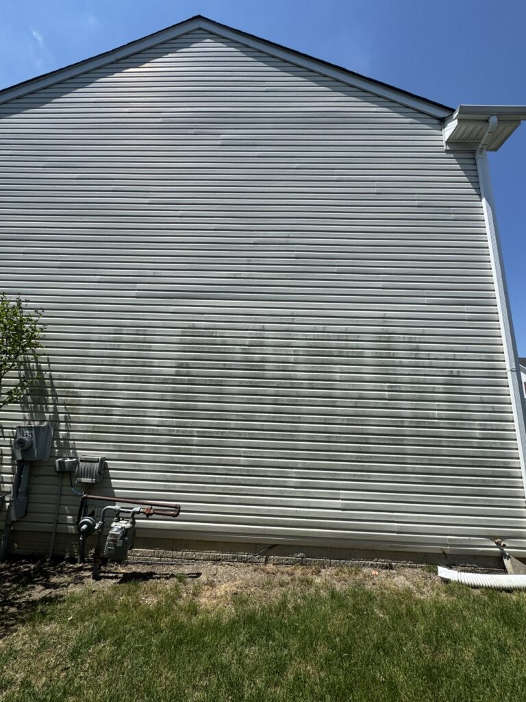 Side of a house with horizontal light-gray metal siding, gutter and downspout, utility meters at the base, and a blue sky above.