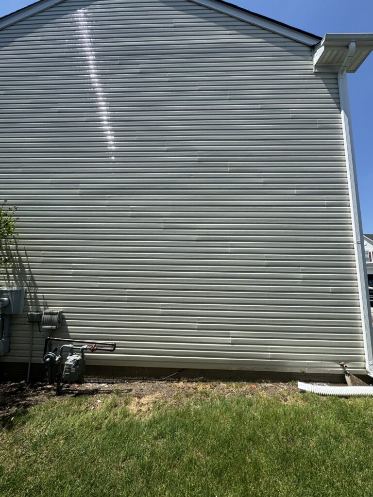 Side of a house with gray horizontal vinyl siding, blue sky above, utility meters and pipes at the base, and a grassy foreground.