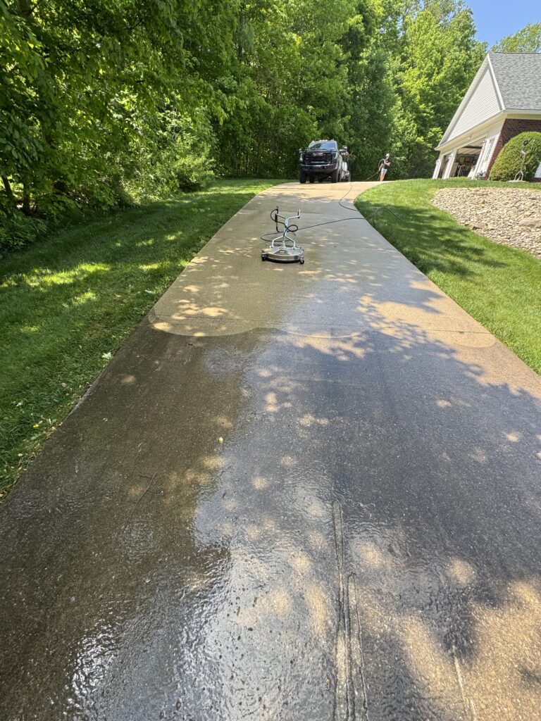 A long driveway being pressure-washed; a user in the background operates a power washer with a dark truck nearby; green lawn and trees frame the scene on the left and a house is on the right.
