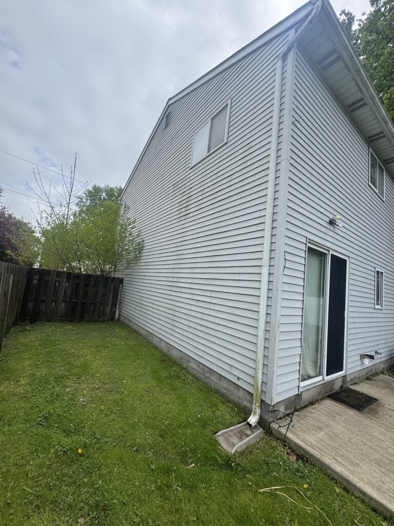 Backyard view of a light-gray siding house with sliding glass door, downspout, and small windows beside a wooden fence under a cloudy sky