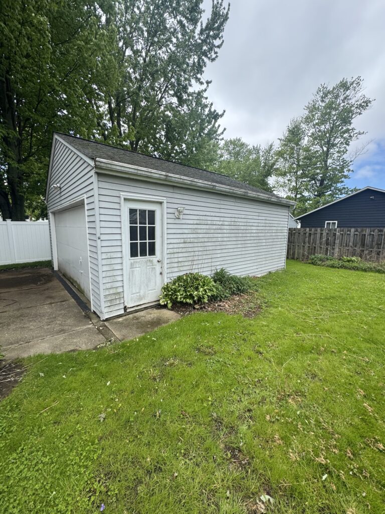 White wooden shed with a door and small window, set in a green yard with tall trees behind it.“