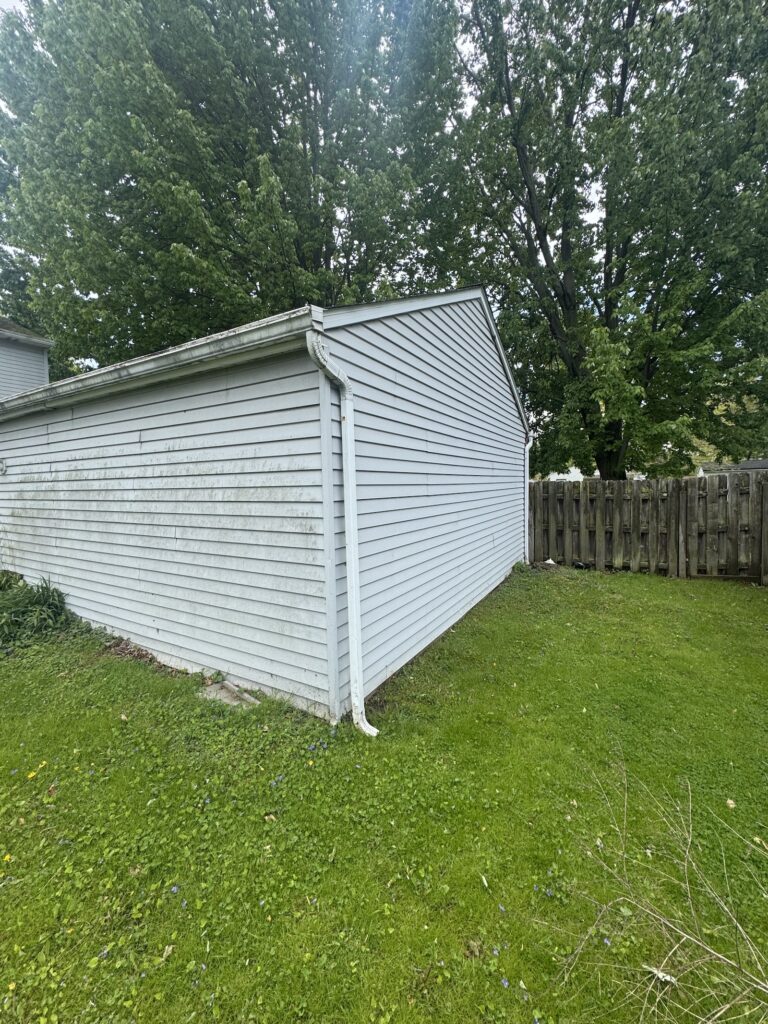 White wooden shed with a sloped roof and rain gutter, located in a green yard with a large tree and wooden fence in the background.