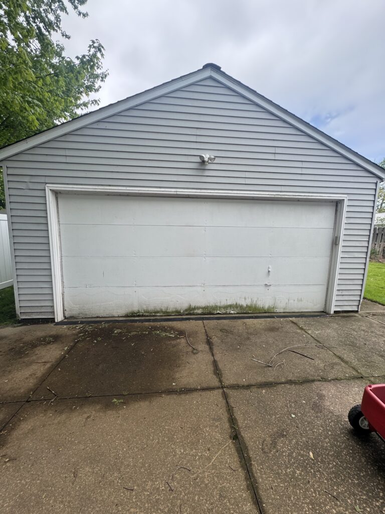 Blue-gray detached garage with a white large roll-up door and a peaked roof under an overcast sky.