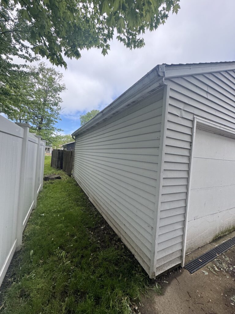 Narrow grassy alley between a white fence and a light-gray shed, with overhanging tree branches and a cloudy sky above.