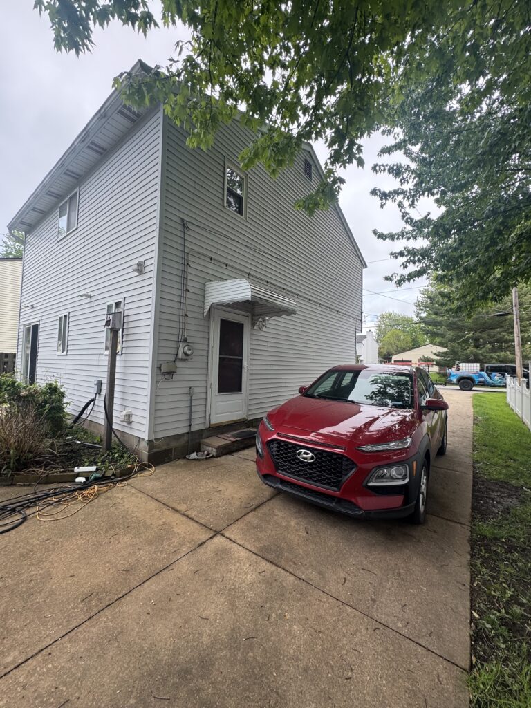 Two-story gray-siding house with a small white awning over the back door and a red SUV parked in the driveway with overhanging tree branches above.
