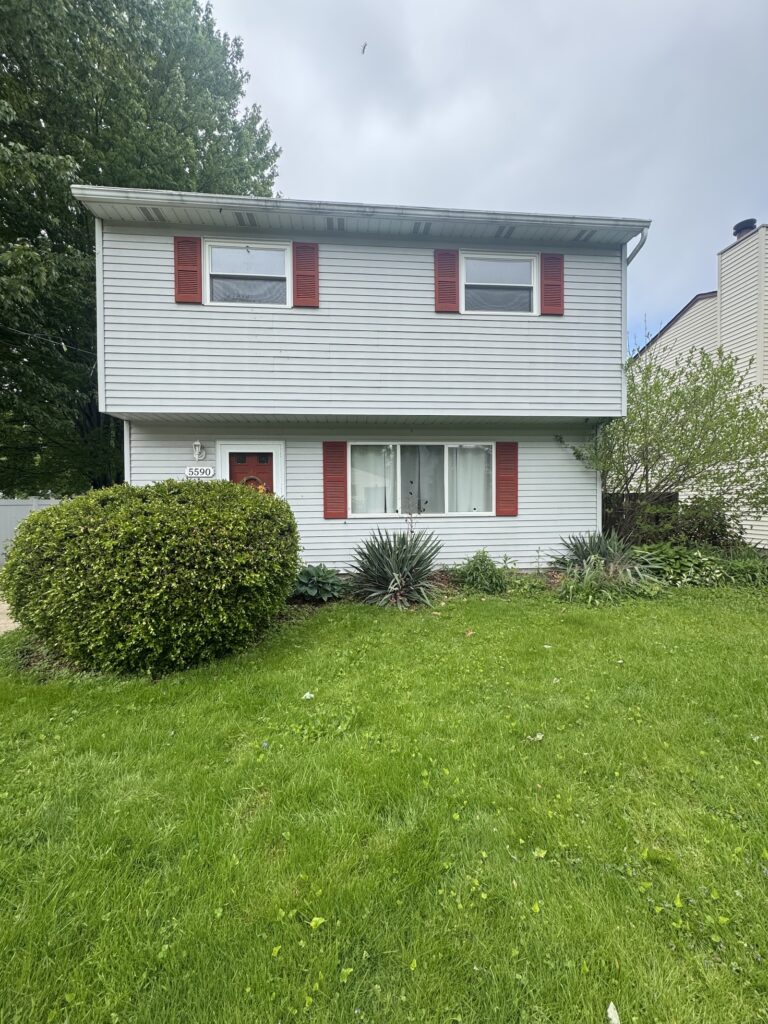 Two-story gray house with red shutters, front yard lawn and trimmed shrubs.