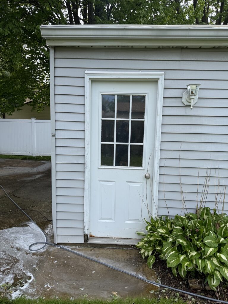White door on a gray siding shed with an outdoor light, surrounded by water pooling on the concrete and a garden hose nearby.
