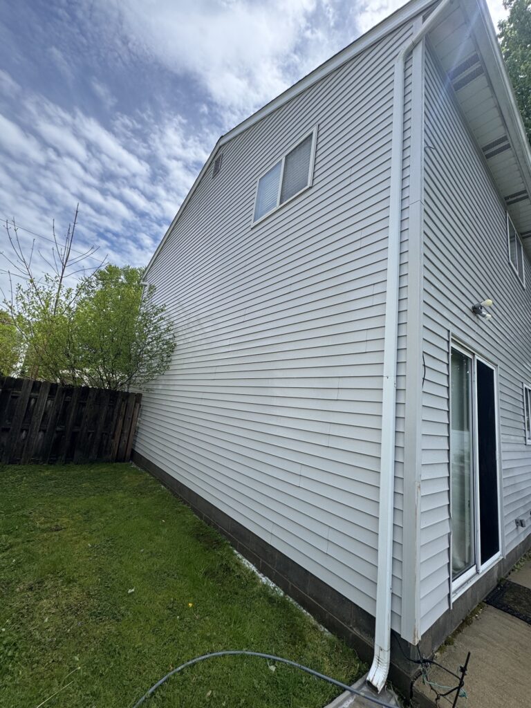 Side view of a light gray house with white siding, gutter downspout, and sliding glass door along a grassy yard under a blue sky.