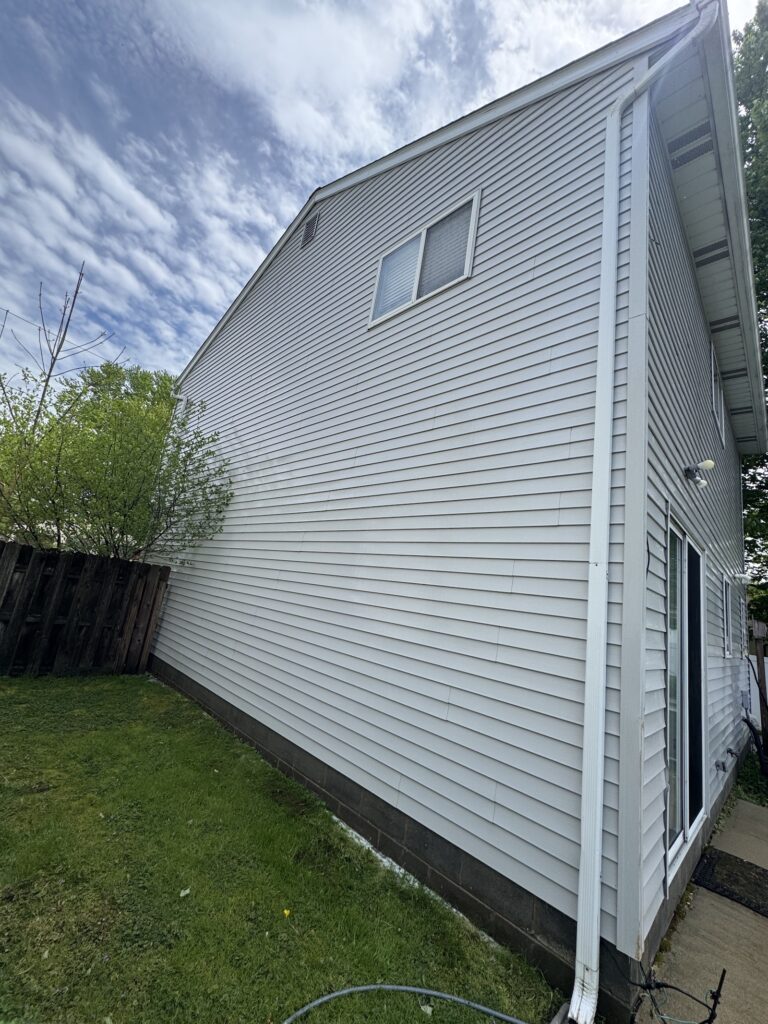 Side of a light-gray vinyl-sided house with a white gutter and downspout, small second-story window, and a green lawn nearby under a blue sky.