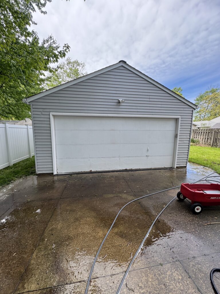 Gray detached garage with a white door, wet concrete driveway, and a red wagon nearby in a residential yard after rainsi