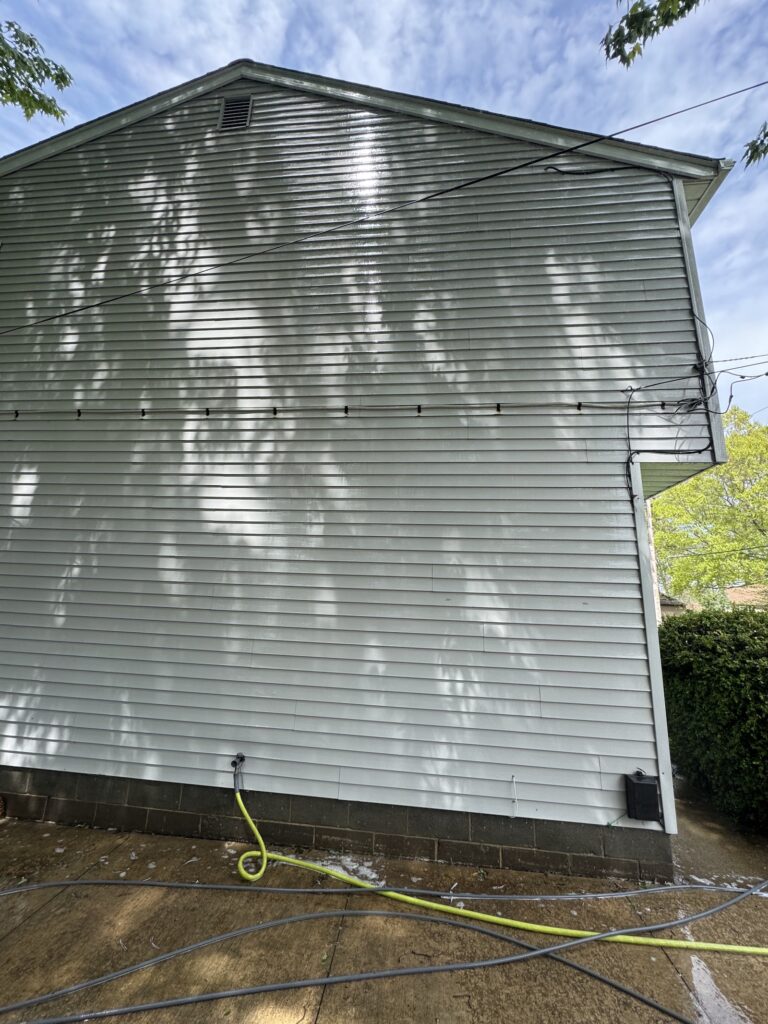 Side of a light-gray siding house with tree shadows cast across it and a few cables running along the wall. A bright yellow hose lies on the wet concrete ground nearby.