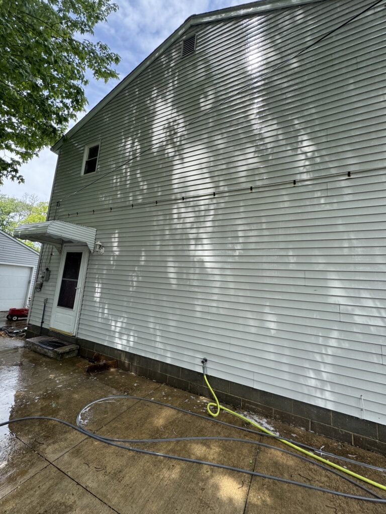 Side of a gray clapboard house with a small window, on a wet concrete yard with hoses lying on the ground and leafy tree shadows.