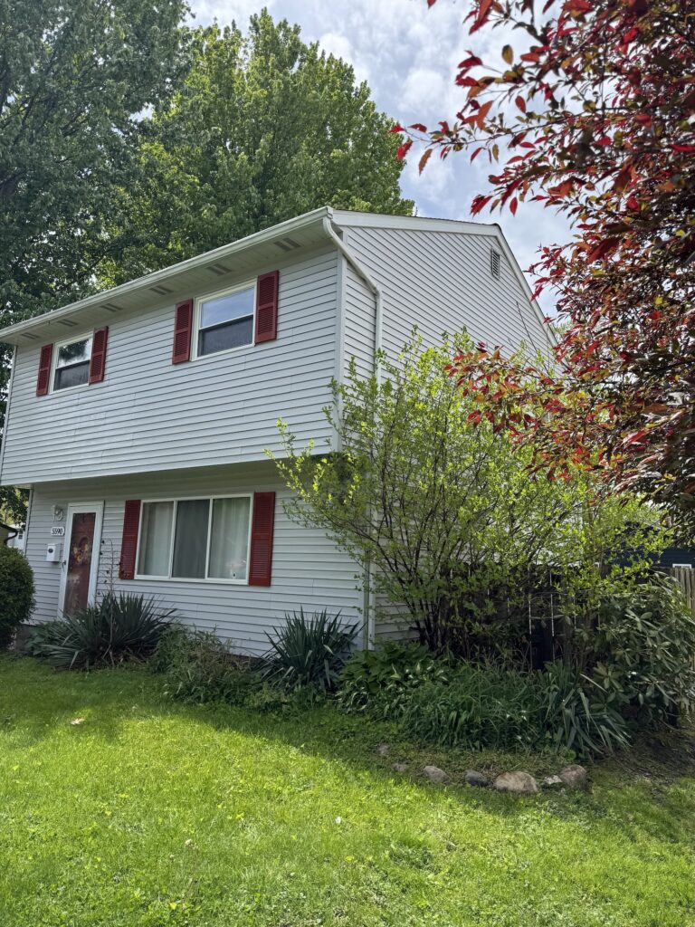 White two-story suburban house with red shutters and large front windows, surrounded by green lawn and trees in a sunny yard.