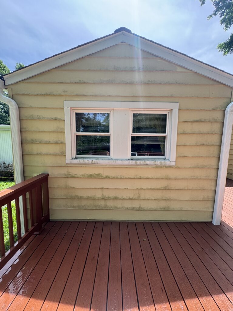 Back exterior wall of a small house with pale yellow siding, white-framed window, and a wooden deck; sun flare across the image.