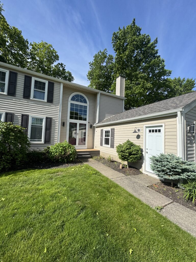 Front of a two-story beige house with black shutters, large arched window, and a chimney, surrounded by a green lawn and shrubs under a blue sky