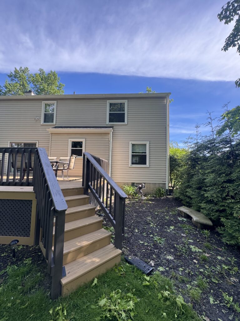 Backyard scene with a wooden deck and stairs, beige house siding, outdoor seating, and a blue sky above.