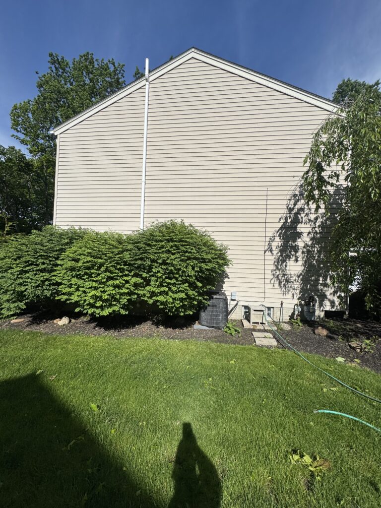 Backyard view of a beige house with white downspout, greenery along the foundation, and a visible outdoor air-conditioner unit; blue sky overhead.