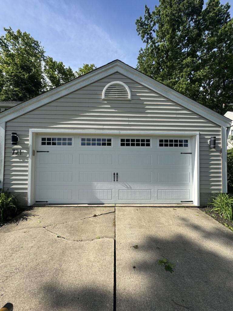 Front view of a beige house garage with a white double door and a decorative arched vent above, surrounded by trees and a concrete driveway.
