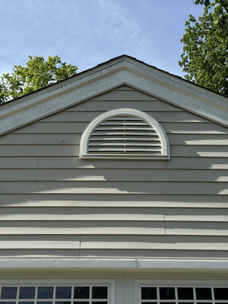 Front of a beige house with a white semicircular vent under the triangular roof, blue sky and green trees in the background.