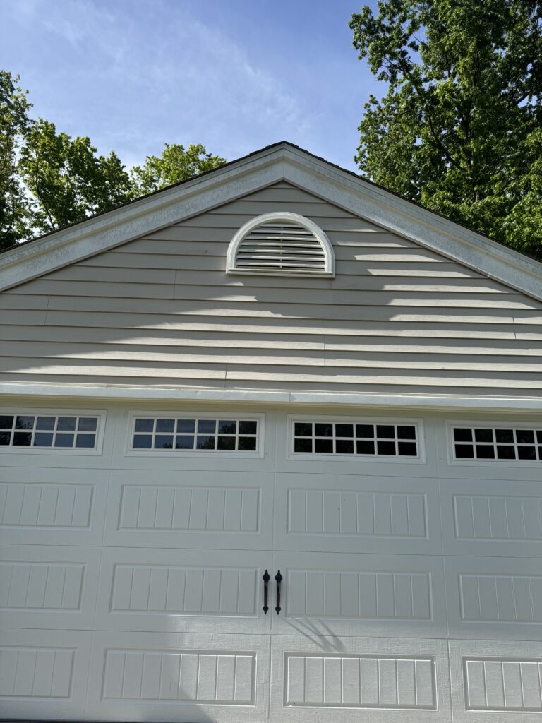 White garage door with two rows of small rectangular windows near the top, beige siding, and a circular white vent centered on the upper wall under the roof peak.