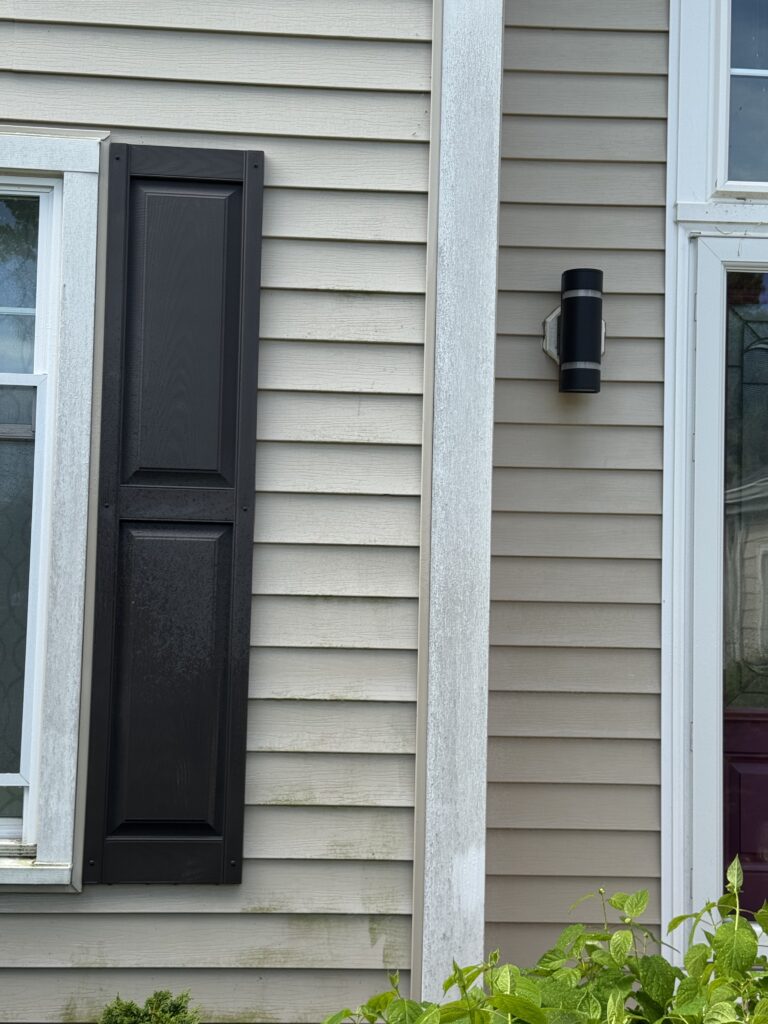 Exterior view of a beige house wall with a black, closed, vertical shutter beside a window and a black cylindrical wall light mounted to the siding; green plants at the bottom.
