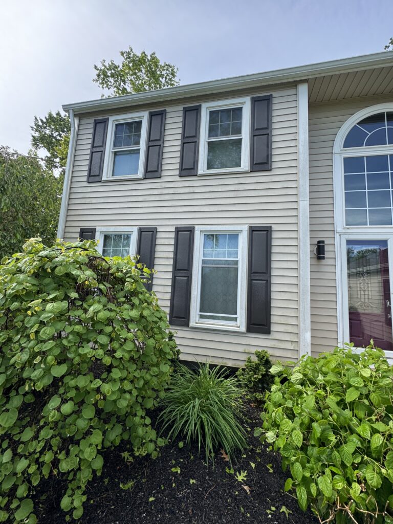 Beige siding house with white-trimmed windows and dark shutters, two stories high, front yard filled with green shrubs and mulch.
