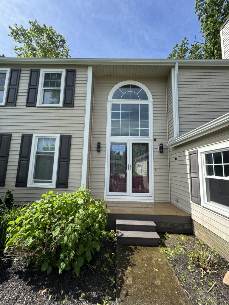 Front entry of a beige house with an arched window above double doors, flanked by black shutters and green shrubbery in front.