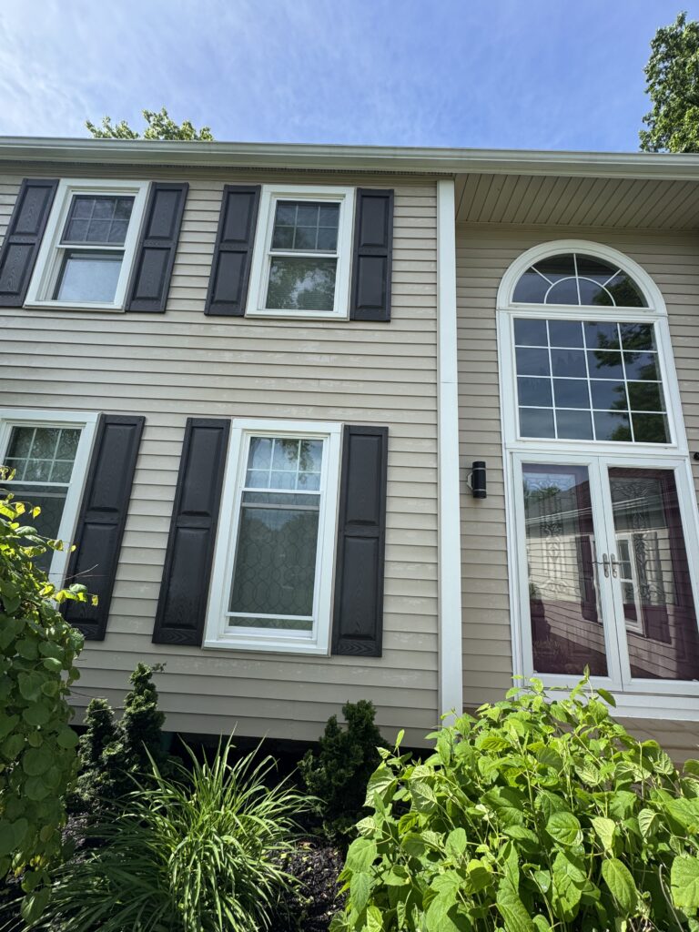 Front of a beige house with white-trimmed windows, dark shutters, and a large arched window to the right, amid garden greenery.