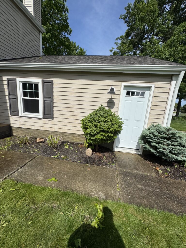 Side view of a beige house wall with a white door, dark shutters, and a wall-mounted light, surrounded by shrubs and a concrete path.
