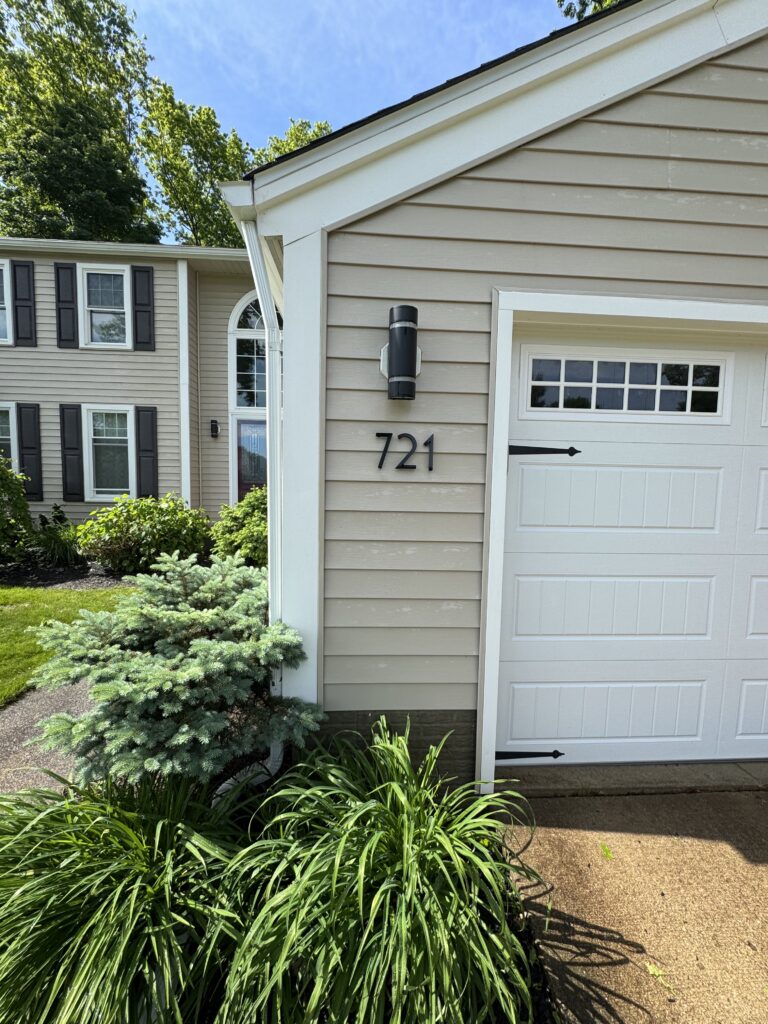 Beige house with white trim and a black exterior light near the address 721, a white garage door, and lush green shrubs in the yard.