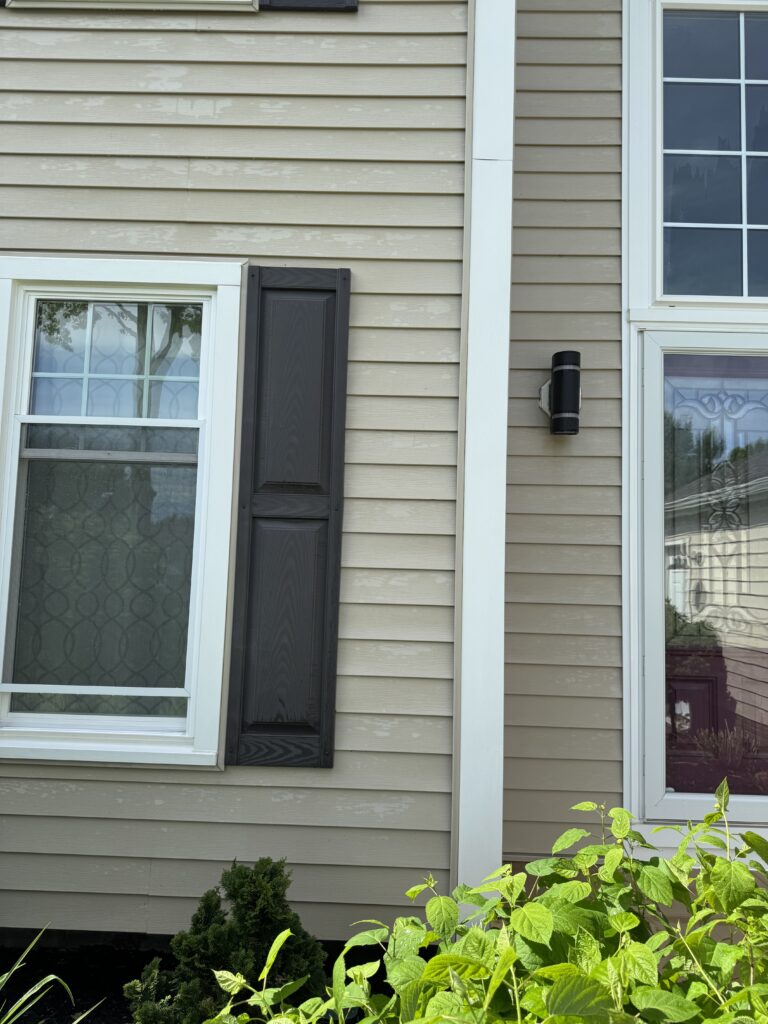 Front of house exterior: beige siding, white window trim, black shutters, and a black wall light with greenery in the foreground.