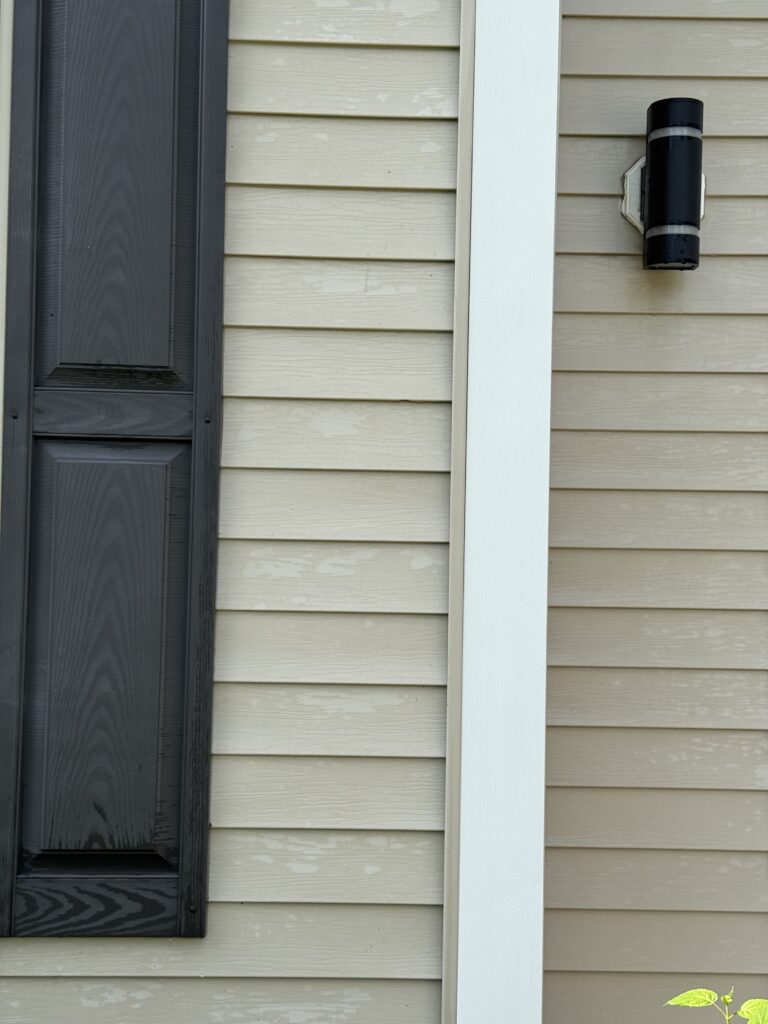 Exterior wall with beige horizontal siding, a dark door frame on the left, and a white vertical trim with a black outdoor wall light on the right.