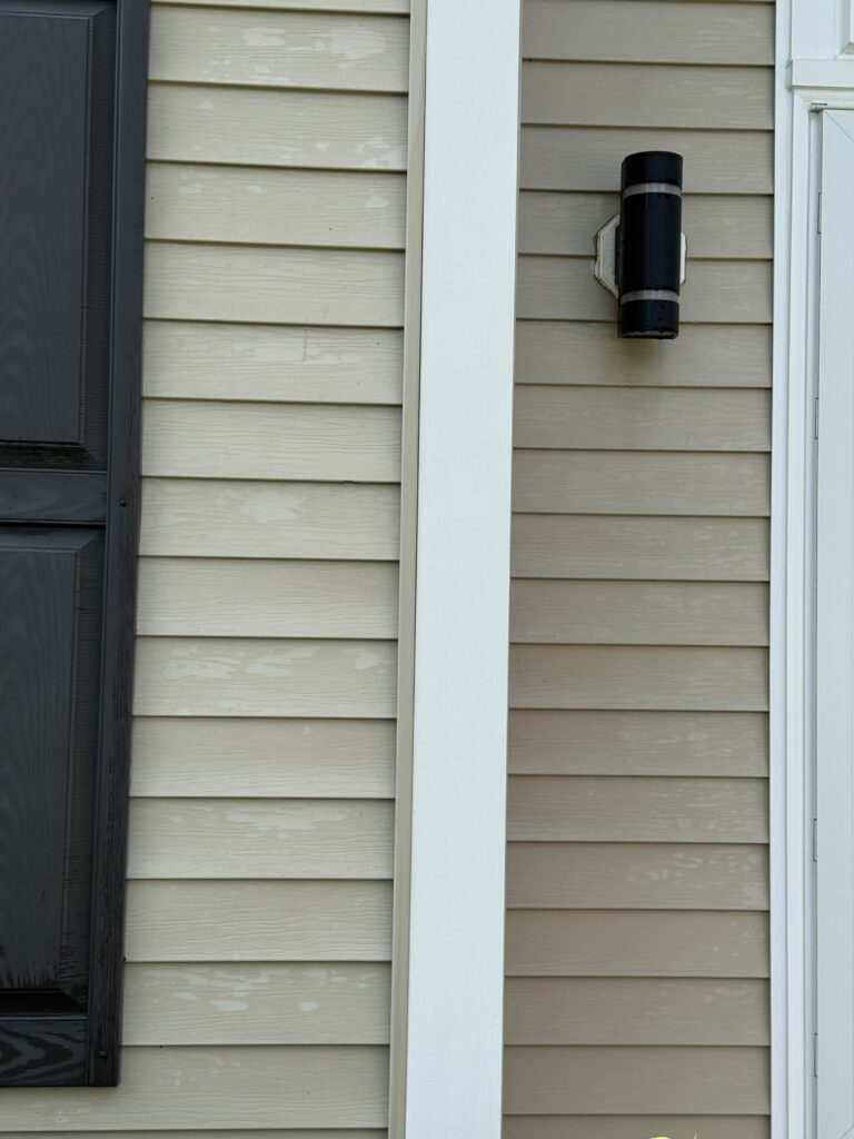 Exterior wall of a house with beige horizontal siding, a vertical white trim beam, and a black wall-mounted outdoor light.