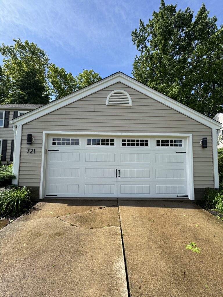 Garage with beige siding and a white multi-panel door, arched vent above, driveway in front, and trees against a blue sky.