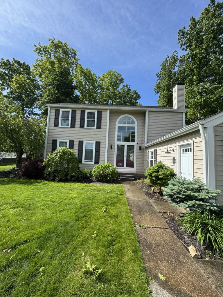 Front view of a beige, two-story house with arched window, white trim, and black shutters, set in a green lawn garden.