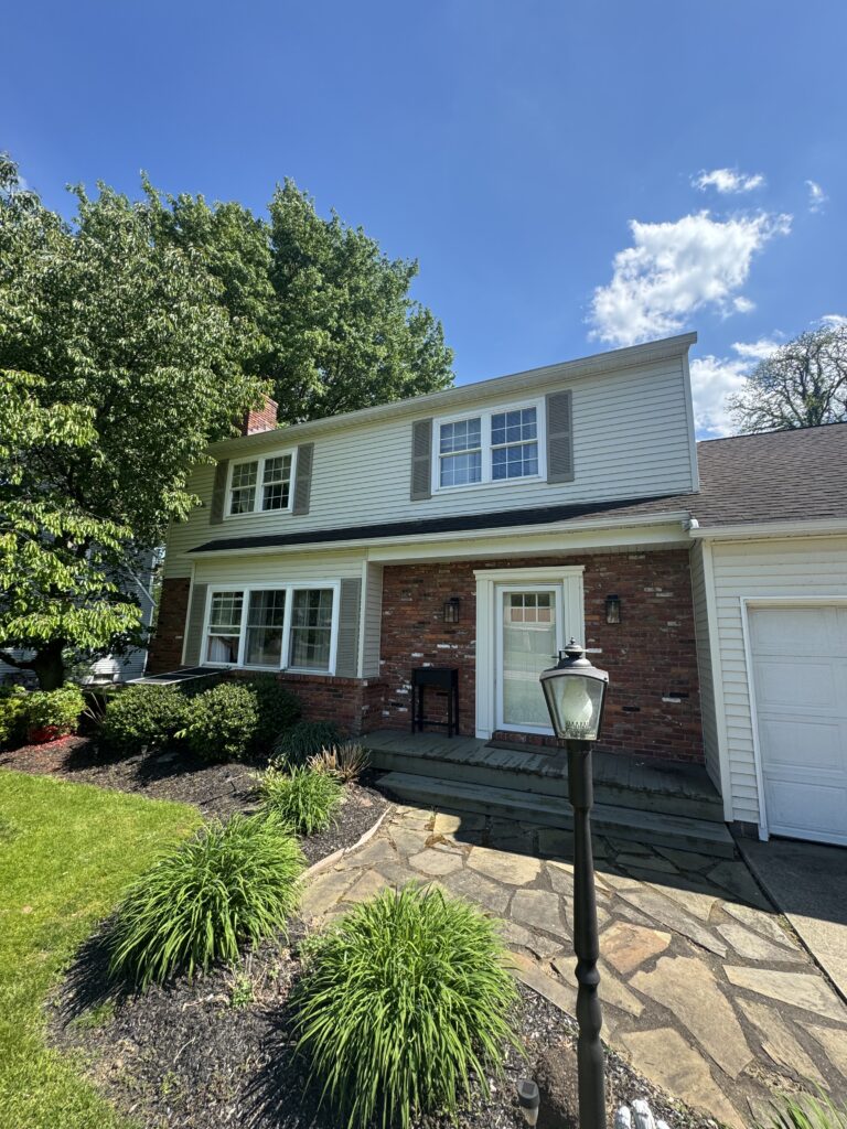 Front of a two-story suburban house with beige siding, brick lower level, two windows with shutters, and a stone pathway.
