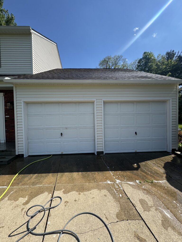 Two-car white garage doors on a beige house with a sloped roof, wet concrete driveway, and colorful hoses lying in the foreground under a bright blue sky.