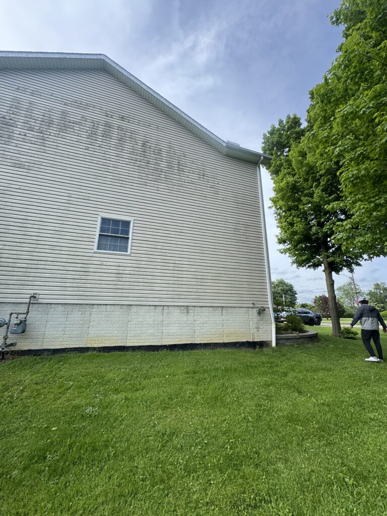 Side view of a beige vinyl-sided building with a curved roofline, small window, and a grassy yard; green tree on the right and a person walking nearby.