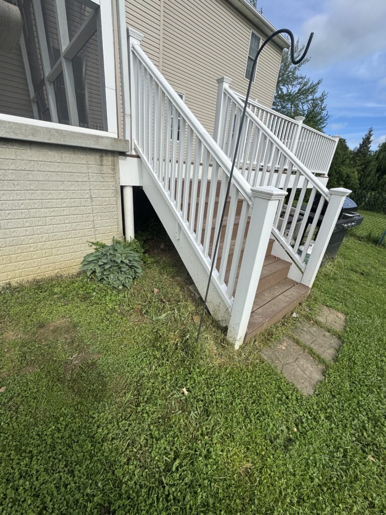 Backyard stairs with white railing leading to a back door, beige siding house, and a black hose looping overhead with a grill on the lawn.