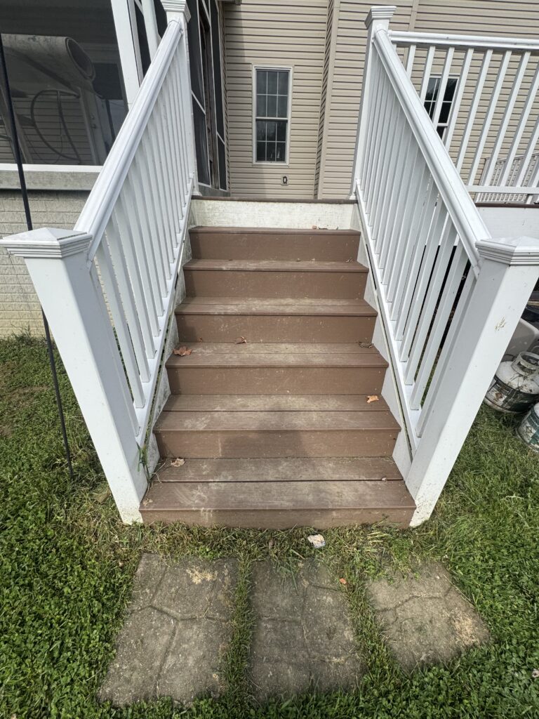Outdoor wooden stairs with white railings lead to a back door, beige siding house in view; grass and a concrete path at ground level.