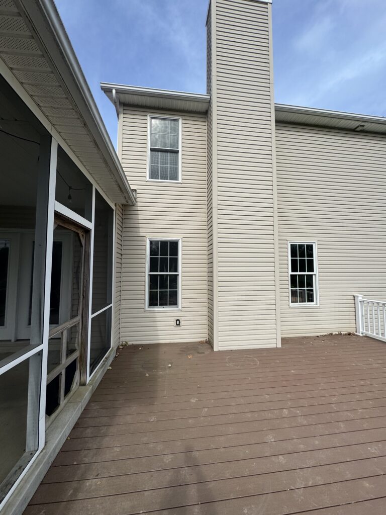 Backyard deck beside a beige two-story house with tall chimney and multiple windows under a blue sky, seen from the patio door area.
