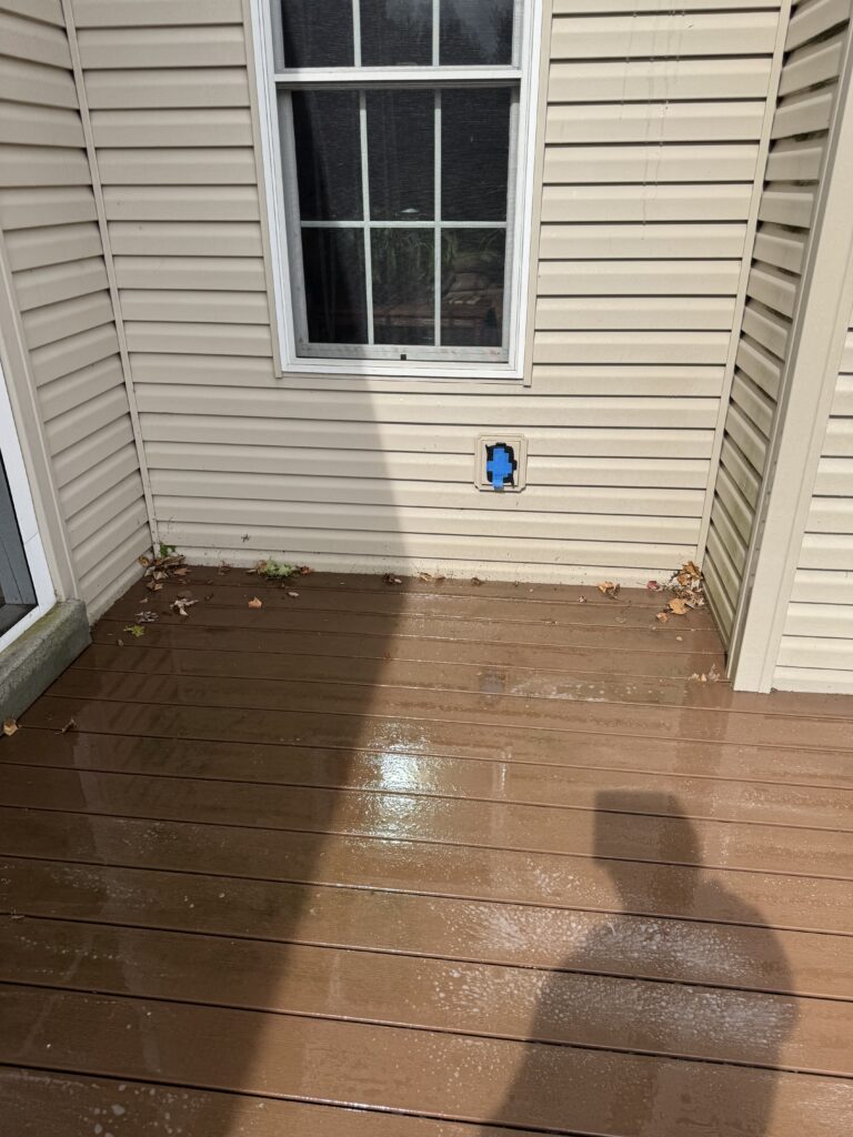 Beige vinyl-siding wall with a white-framed window; a small square wall opening has a blue taped cover, on a wet brown wooden deck with scattered leaves nearby.